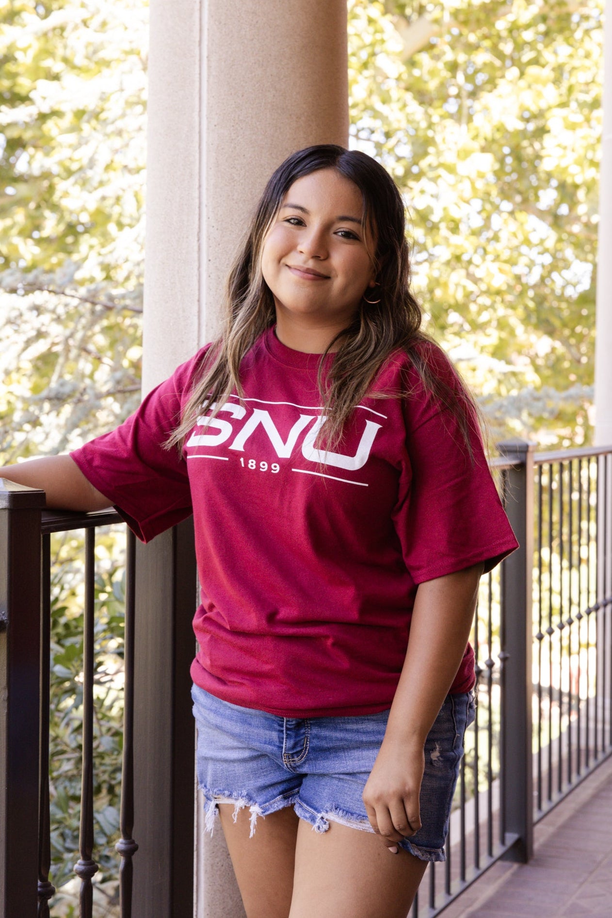 Person wearing a red t-shirt with 'SNU' on it, standing on a balcony with trees in the background